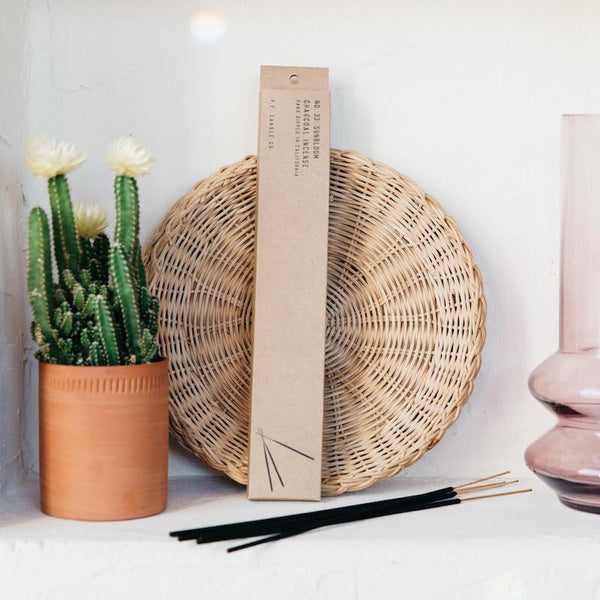 PF Candle Co Sunbloom incense package leaning against a white wall, between a potted cactus and pink glass with incense sticks laying in front